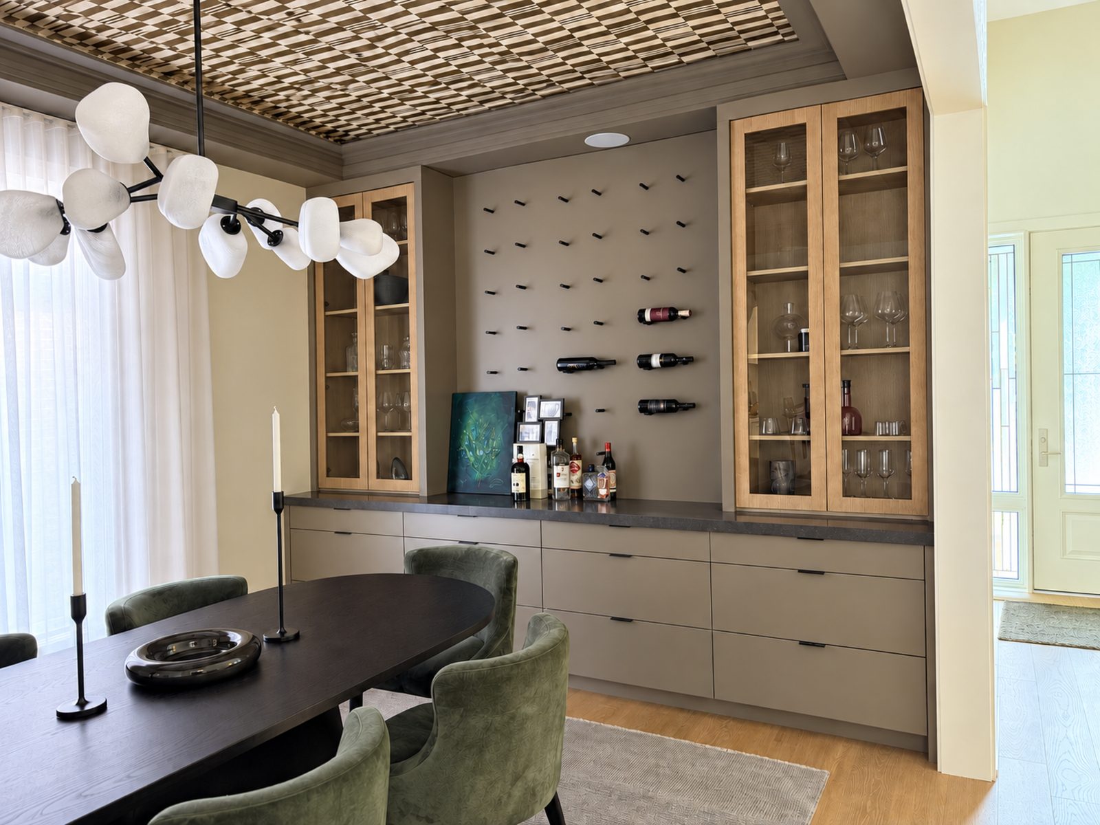 Ajax house dining room with a dark oval table, green velvet chairs and candlesticks, beneath a checkerboard ceiling detail, facing a built-in servery of white oak glass-front cabinets, a stone counter and a wall-mounted wine rack