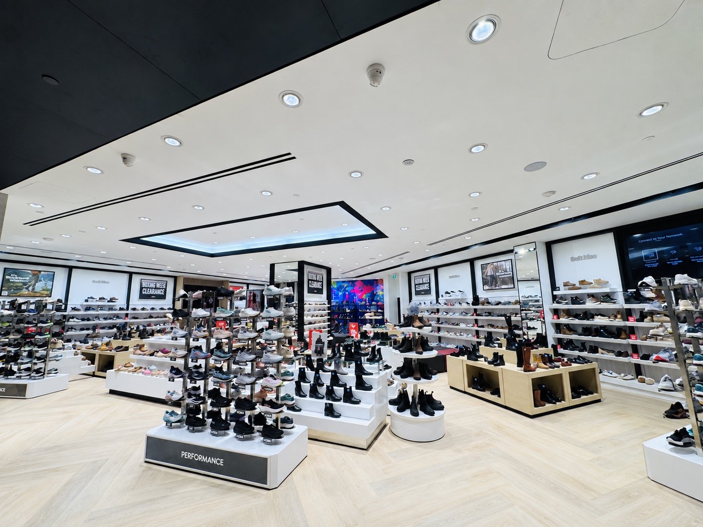 SoftMoc retail store interior at Square One with tiered white display tables of footwear, a PERFORMANCE feature table, wall shelving and a mirrored feature wall