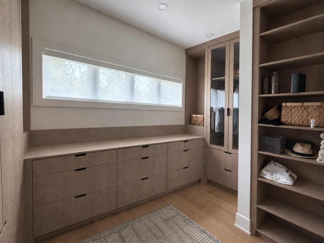 White oak dressing room with a long drawer run under a clerestory window, a glass-front wardrobe in the corner, and open shelving alongside