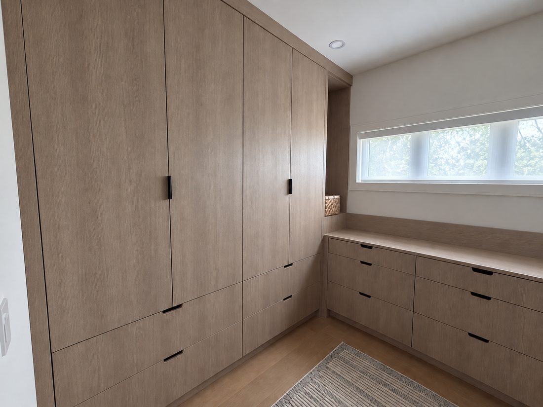 Floor-to-ceiling white oak wardrobe wall with recessed black edge pulls, turning the corner into a fitted drawer run beneath a clerestory window